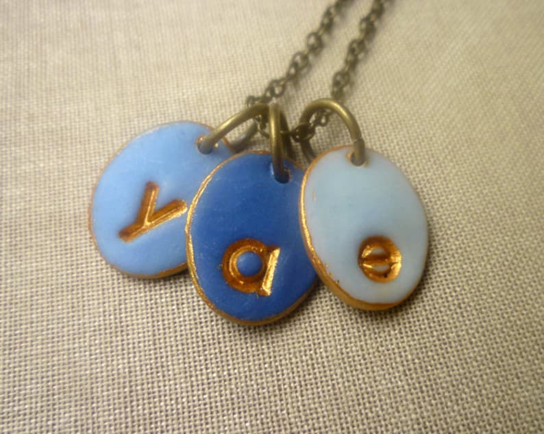 May include: A gold chain necklace with three blue and white ceramic pendants. Each pendant has a gold letter on it, spelling out the word "Ava".