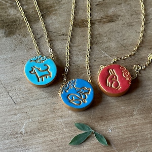 May include: Three gold chain necklaces with blue and red ceramic pendants. Each pendant features a different animal design: a dog, a squirrel, and a rabbit. The pendants have a gold-colored outline and intricate details.
