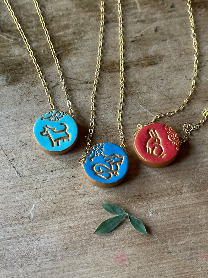 May include: Three gold chain necklaces with blue and red ceramic pendants. Each pendant features a different animal design: a dog, a squirrel, and a rabbit. The pendants have a gold-colored outline and intricate details.