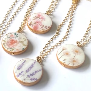 May include: Four gold chain necklaces with round pendants featuring different floral designs. The pendants are white with watercolour floral illustrations of pink flowers, lavender, and white daisies.