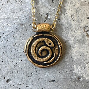 May include: A gold-colored necklace with a circular pendant featuring a black and gold snake design. The snake is coiled in a spiral pattern. The necklace has a delicate gold chain and clasp.