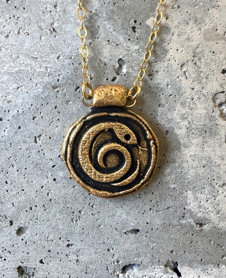 May include: A gold-colored necklace with a circular pendant featuring a black and gold snake design. The snake is coiled in a spiral pattern. The necklace has a delicate gold chain and clasp.
