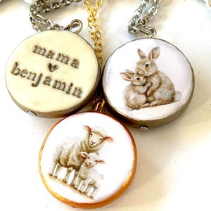 Puede incluir: Tres collares con colgantes redondos. Un colgante tiene las palabras "mama ♥ benjamin" en letras en relieve. Los otros dos colgantes presentan ilustraciones de un conejo madre e hijo y una oveja madre con un cordero. Los colgantes tienen cadenas doradas y plateadas.