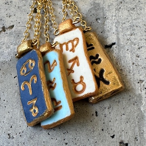 May include: Four rectangular pendant necklaces with gold chains. Each pendant features a different zodiac sign symbol in gold against a blue, light blue, white, and gold background. The pendants are arranged in a row.