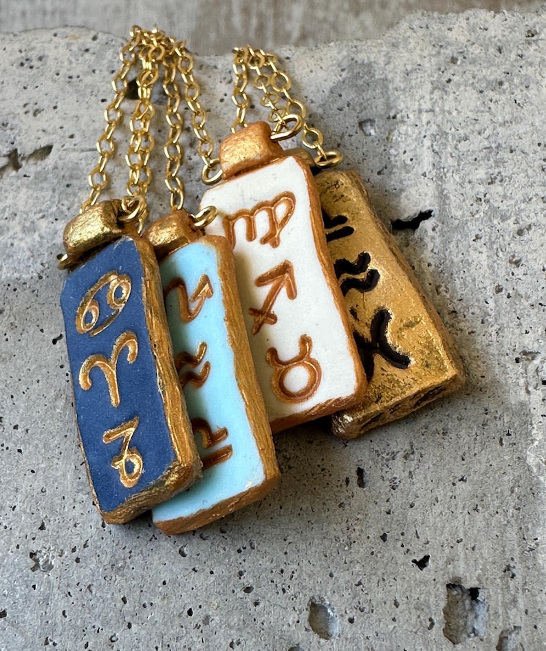 May include: Four rectangular pendant necklaces with gold chains. Each pendant features a different zodiac sign symbol in gold against a blue, light blue, white, and gold background. The pendants are arranged in a row.