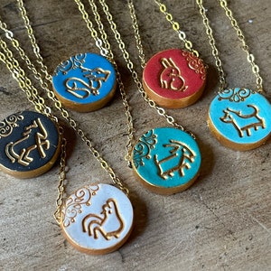 May include: Six gold necklaces with round ceramic pendants featuring different animal designs. The pendants are decorated with gold paint and white floral patterns. The animals include a horse, a rooster, a dog, a rabbit, a goat, and a fox.