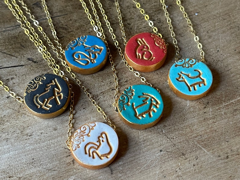 May include: Six gold necklaces with round ceramic pendants featuring different animal designs. The pendants are decorated with gold paint and white floral patterns. The animals include a horse, a rooster, a dog, a rabbit, a goat, and a fox.