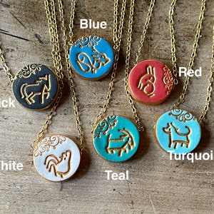 May include: Six ceramic pendants on gold chains, each with a different animal design and color. The pendants are black, white, teal, blue, red, and turquoise. The colors are labeled below each pendant.
