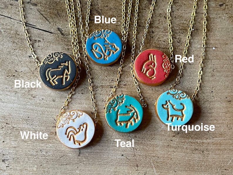 May include: Six ceramic pendants on gold chains, each with a different animal design and color. The pendants are black, white, teal, blue, red, and turquoise. The colors are labeled below each pendant.