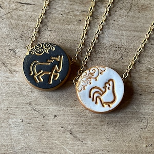 May include: Two gold chain necklaces with round ceramic pendants. One pendant is black with a gold horse design and the other is white with a gold rooster design.