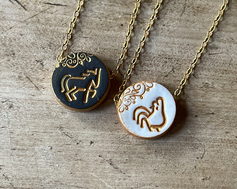May include: Two gold chain necklaces with round ceramic pendants. One pendant is black with a gold horse design and the other is white with a gold rooster design.