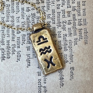 May include: A gold-colored pendant necklace featuring a rectangular charm with black zodiac symbols. The charm is suspended from a delicate gold chain. The background is an aged paper with German text.