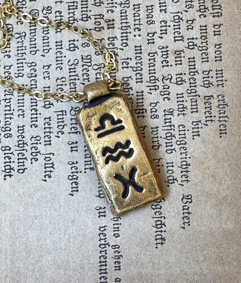 May include: A gold-colored pendant necklace featuring a rectangular charm with black zodiac symbols. The charm is suspended from a delicate gold chain. The background is an aged paper with German text.