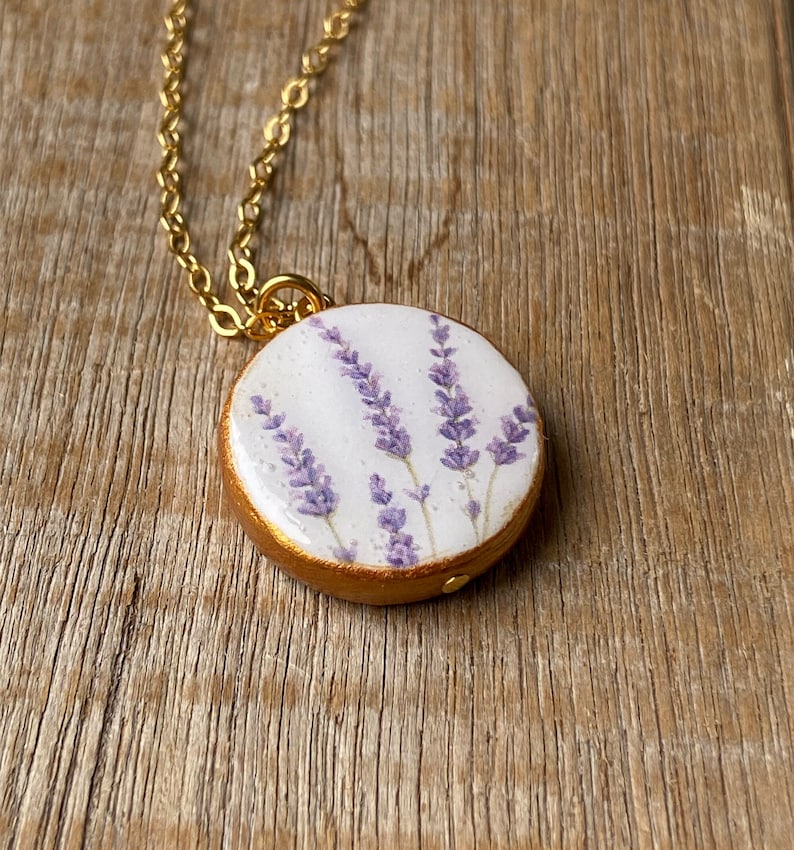 May include: A gold chain necklace with a round pendant featuring a pressed lavender flower design on a white background.