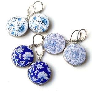 May include: Several pairs of round earrings featuring blue and white floral patterns. The earrings have silver-coloured lever-back fastenings. The floral designs vary, with different botanical styles. The earrings are presented against a white backdrop.