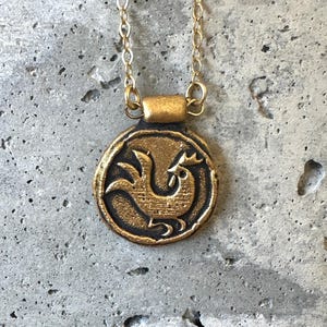May include: A gold-colored pendant necklace featuring a circular charm with a rooster design. The pendant hangs from a delicate gold chain. The rooster is depicted in a stylized, vintage style. The necklace is displayed against a textured, gray background.