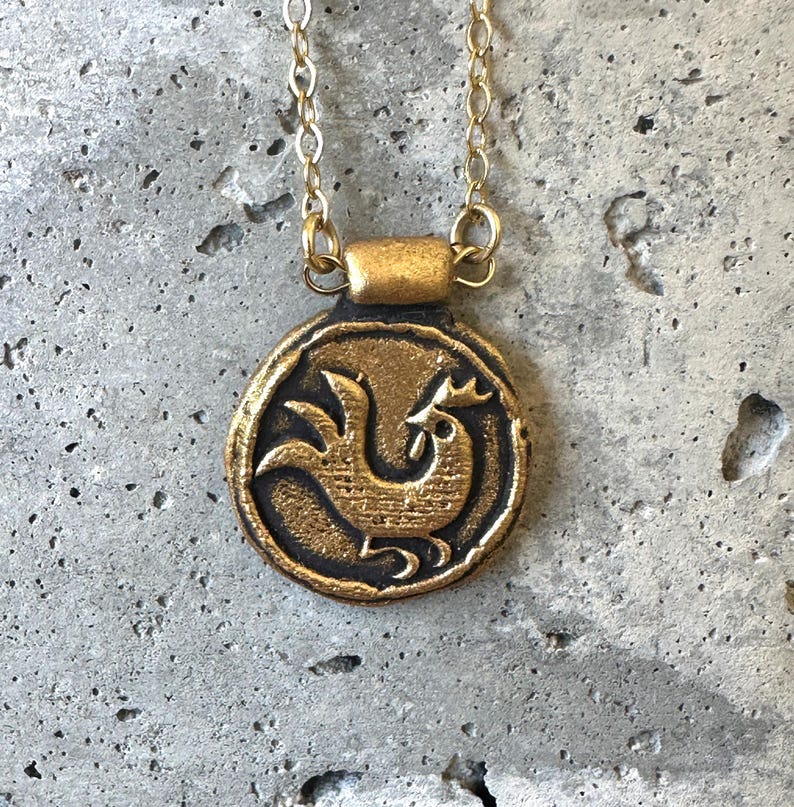 May include: A gold-colored pendant necklace featuring a circular charm with a rooster design. The pendant hangs from a delicate gold chain. The rooster is depicted in a stylized, vintage style. The necklace is displayed against a textured, gray background.