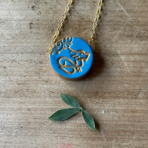 May include: A gold chain necklace with a blue circular pendant featuring a gold design. The pendant has a floral pattern and the word "Om" in gold lettering.