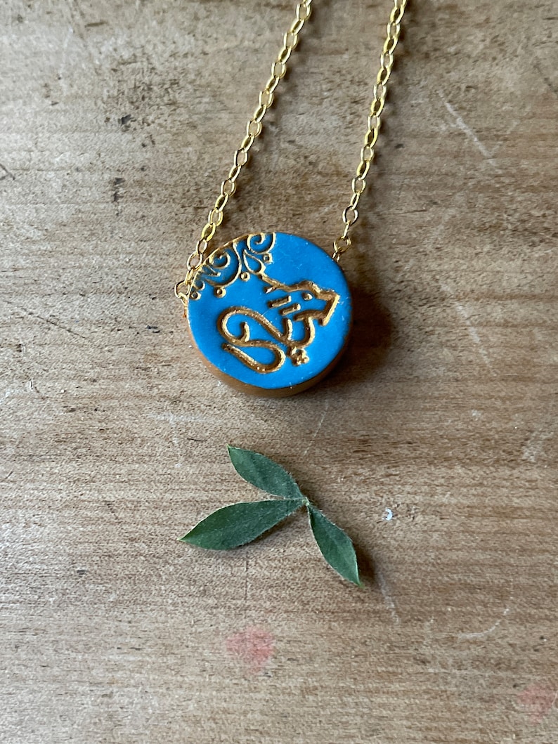 May include: A gold chain necklace with a blue circular pendant featuring a gold design. The pendant has a floral pattern and the word "Om" in gold lettering.