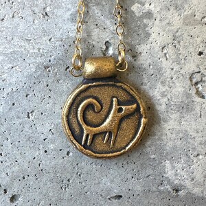 May include: A gold-colored pendant necklace featuring a circular charm with a stylized dog design. The dog is depicted in a simple, linear style. The necklace has a delicate gold chain.
