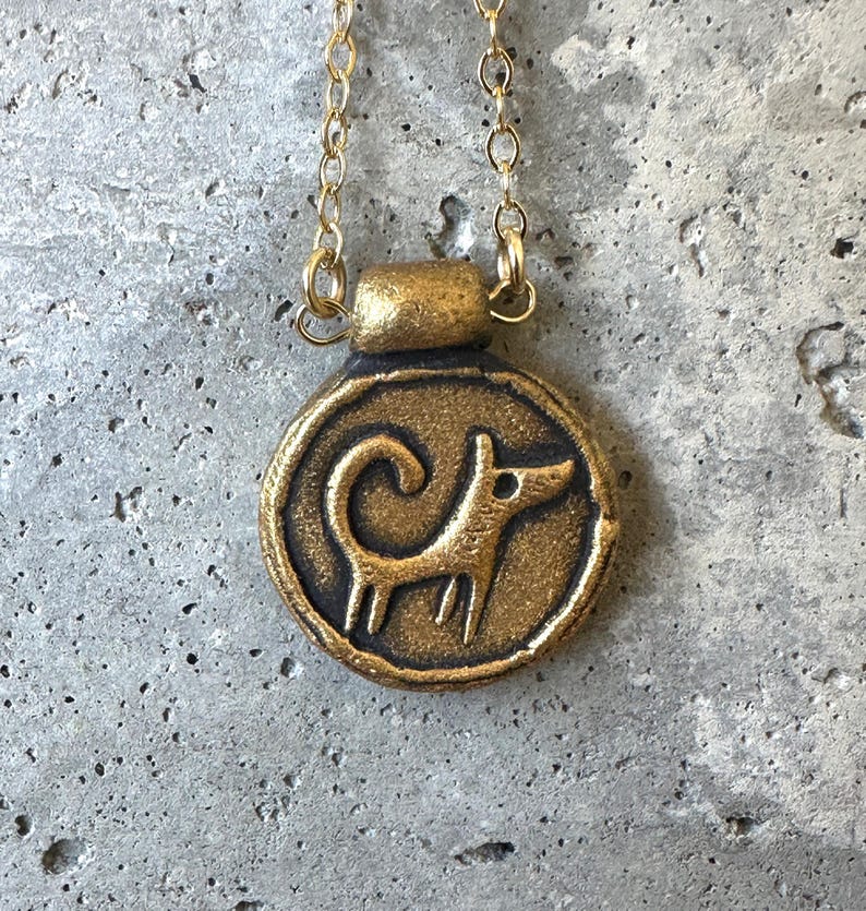 May include: A gold-colored pendant necklace featuring a circular charm with a stylized dog design. The dog is depicted in a simple, linear style. The necklace has a delicate gold chain.
