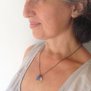 May include: A woman wearing a light gray tank top with a brown chain necklace with two small oval pendants. One pendant is blue with a gold dot and the other is gold with a blue dot.