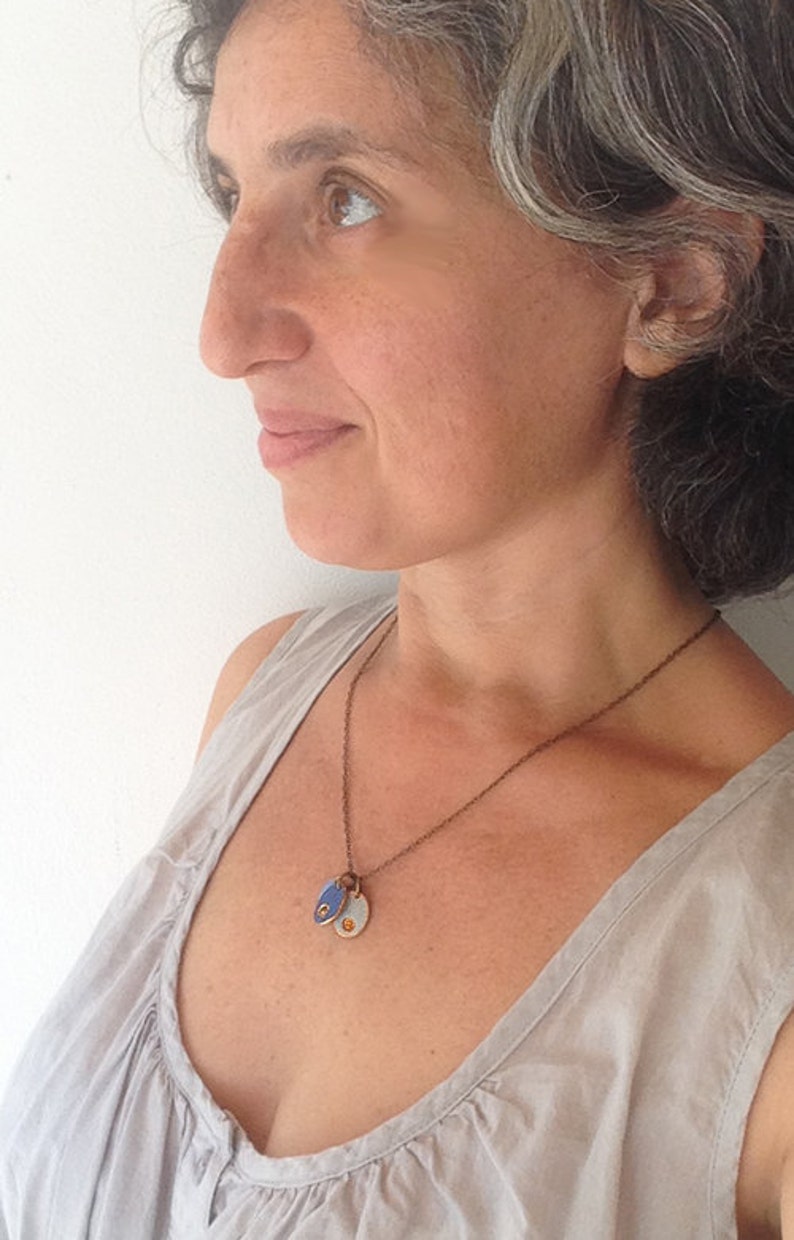 May include: A woman wearing a light gray tank top with a brown chain necklace with two small oval pendants. One pendant is blue with a gold dot and the other is gold with a blue dot.