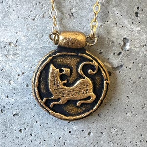 May include: A gold-colored pendant necklace with a circular, black and gold medallion. The medallion features a detailed relief of a stylized feline figure. The necklace chain is also gold-colored, and the pendant is suspended against a gray background.