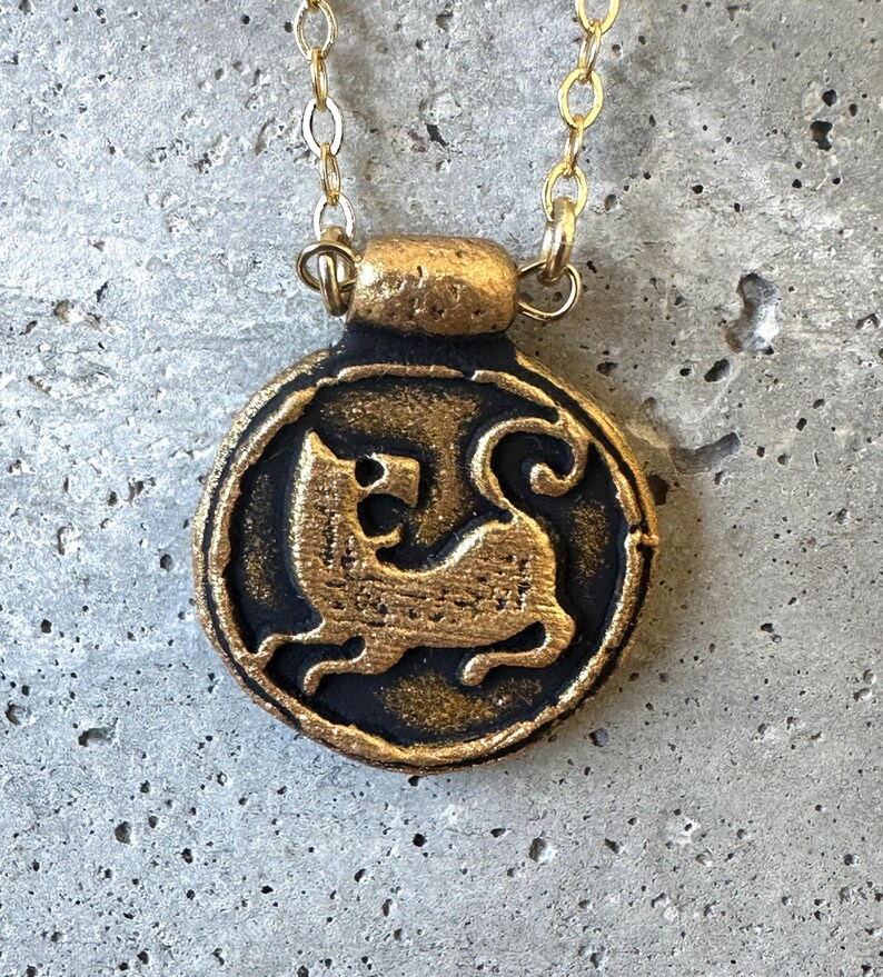 May include: A gold-colored pendant necklace with a circular, black and gold medallion. The medallion features a detailed relief of a stylized feline figure. The necklace chain is also gold-colored, and the pendant is suspended against a gray background.