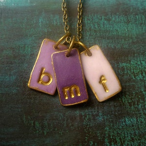 May include: A gold chain necklace with three purple and pink ceramic tags. The tags have the letters "b", "m", and "f" written in gold.