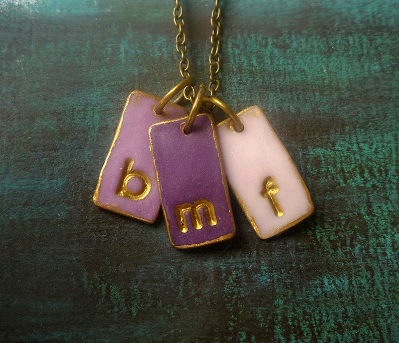 May include: A gold chain necklace with three purple and pink ceramic tags. The tags have the letters "b", "m", and "f" written in gold.