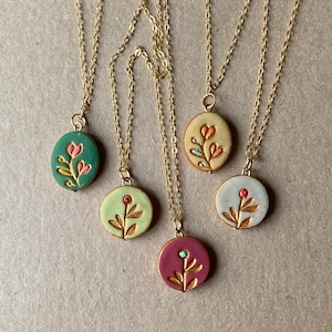 Puede incluir: Cinco collares de cadena dorada con colgantes de cerámica de diferentes colores. Cada colgante presenta un diseño floral sencillo con un contorno dorado.