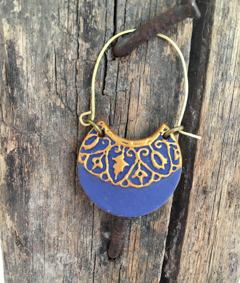 May include: A pair of gold hoop earrings with a blue and gold ceramic crescent moon pendant. The pendant features a gold filigree design.