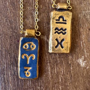 May include: Two gold-colored necklaces with rectangular pendants. One pendant is navy blue with gold zodiac symbols, and the other is gold with black zodiac symbols. The necklaces hang on a gold chain.
