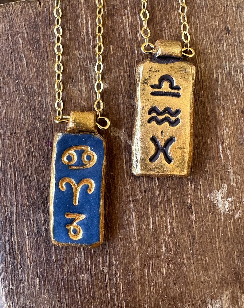 May include: Two gold-colored necklaces with rectangular pendants. One pendant is navy blue with gold zodiac symbols, and the other is gold with black zodiac symbols. The necklaces hang on a gold chain.