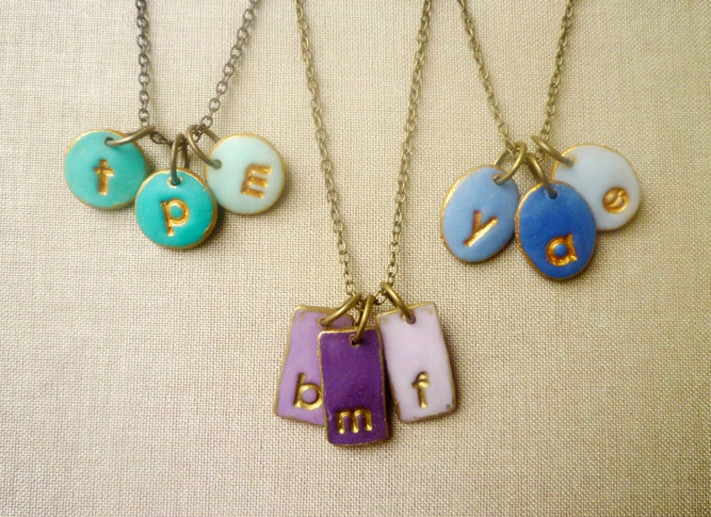 May include: Three gold necklaces with different colored ceramic charms. The first necklace has turquoise charms with the letters "t", "p", and "e". The second necklace has blue charms with the letters "y", "a", and a blank charm. The third necklace has three rectangular purple charms with the letters "b", "m", and "f".