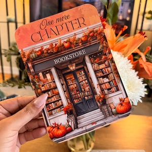 May include: A rectangular card with the text "One more CHAPTER" and "BOOKSTORE" printed on it. The card features an illustration of a bookstore decorated with pumpkins and fall foliage. The card is held in front of a blurred background.