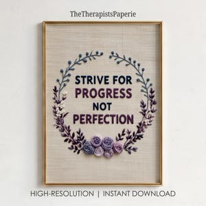 Pode incluir: Uma peça de bordado emoldurada sobre tecido bege com uma coroa de flores em tons de roxo, azul e branco. O texto no centro diz: STRIVE FOR PROGRESS NOT PERFECTION. O design está contido numa moldura fina de metal de cor dourada.