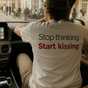 Peut inclure: Un t-shirt blanc avec le texte "Stop thinking Start kissing" en lettres noires et rouges. La personne portant le t-shirt est à l'intérieur d'une voiture, avec une vue sur une scène de rue à travers le pare-brise.