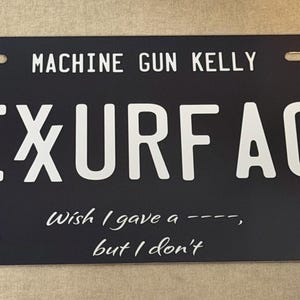 May include: A black rectangular number plate featuring the white text MACHINE GUN KELLY and FIXURFACE. It includes a razor blade graphic, a profile illustration, the text Wish I gave a ----, but I don't, a red Lace Up logo, and a yellow MGK CLEVELAND tag.