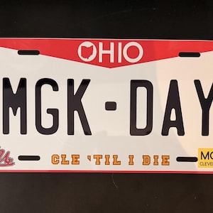 Puede incluir: Una placa de matrícula rectangular con fondo blanco y bordes rojos. El texto MGK-DAY está impreso en letras negras grandes. La parte superior presenta la palabra OHIO con el contorno del estado. El texto incluye CLE 'TIL I DIE y un pequeño gráfico de calavera.