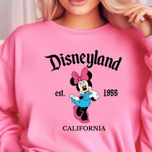 Puede incluir: Sudadera rosa con el logo de Disneyland y el personaje de Minnie Mouse. El dise&ntilde;o incluye el texto "est. 1955" y "CALIFORNIA". Minnie Mouse lleva un vestido azul y un lazo rosa.
