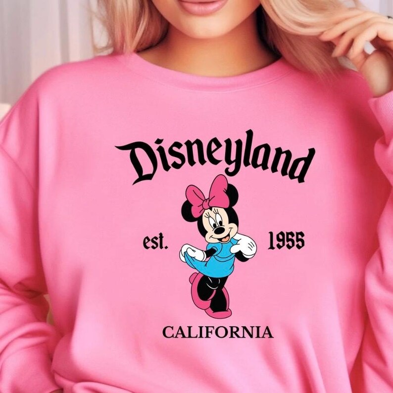 Puede incluir: Sudadera rosa con el logo de Disneyland y el personaje de Minnie Mouse. El dise&ntilde;o incluye el texto "est. 1955" y "CALIFORNIA". Minnie Mouse lleva un vestido azul y un lazo rosa.