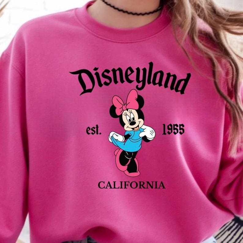 Puede incluir: Una sudadera magenta con el logo de Disneyland en escritura negra, con "est. 1955" y "CALIFORNIA" debajo. Una Minnie Mouse de dibujos animados con un vestido azul y un lazo rosa est&aacute; en el centro.