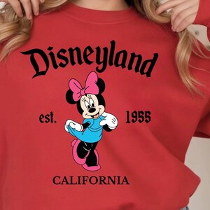 Puede incluir: Una sudadera roja con un gr&aacute;fico de Minnie Mouse en negro, rosa y azul. La palabra "Disneyland" est&aacute; arqueada en la parte superior, con "est. 1955 CALIFORNIA" debajo. La sudadera es de estilo casual.