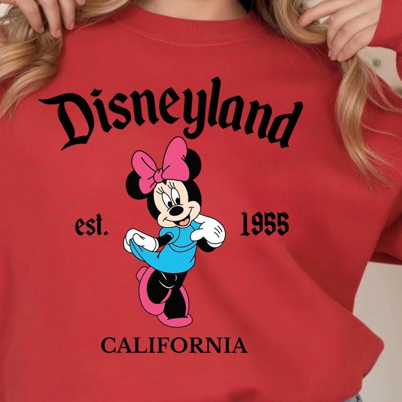 Puede incluir: Una sudadera roja con un gr&aacute;fico de Minnie Mouse en negro, rosa y azul. La palabra "Disneyland" est&aacute; arqueada en la parte superior, con "est. 1955 CALIFORNIA" debajo. La sudadera es de estilo casual.