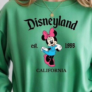 Puede incluir: Sudadera verde con la palabra "Disneyland" en fuente cursiva, y "est. 1955" debajo. Una Minnie Mouse de dibujos animados con un vestido azul y un lazo rosa est&aacute; en el centro. La palabra "CALIFORNIA" est&aacute; en la parte inferior.