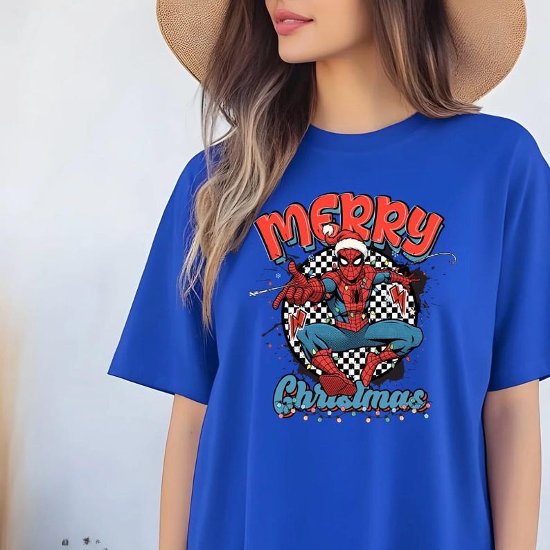 Puede incluir: Camiseta azul real con un gr&aacute;fico de Spider-Man con un gorro de Pap&aacute; Noel. Las palabras "Merry Christmas" est&aacute;n arriba y abajo del personaje en rojo y verde. El dise&ntilde;o est&aacute; sobre un c&iacute;rculo a cuadros blanco y negro.
