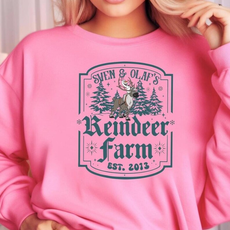 May include: Pink sweatshirt with a graphic featuring the text "Sven & Olaf's Reindeer Farm EST. 2013." The design includes a reindeer, pine trees, and decorative elements in teal and white. The sweatshirt is a solid pink color.