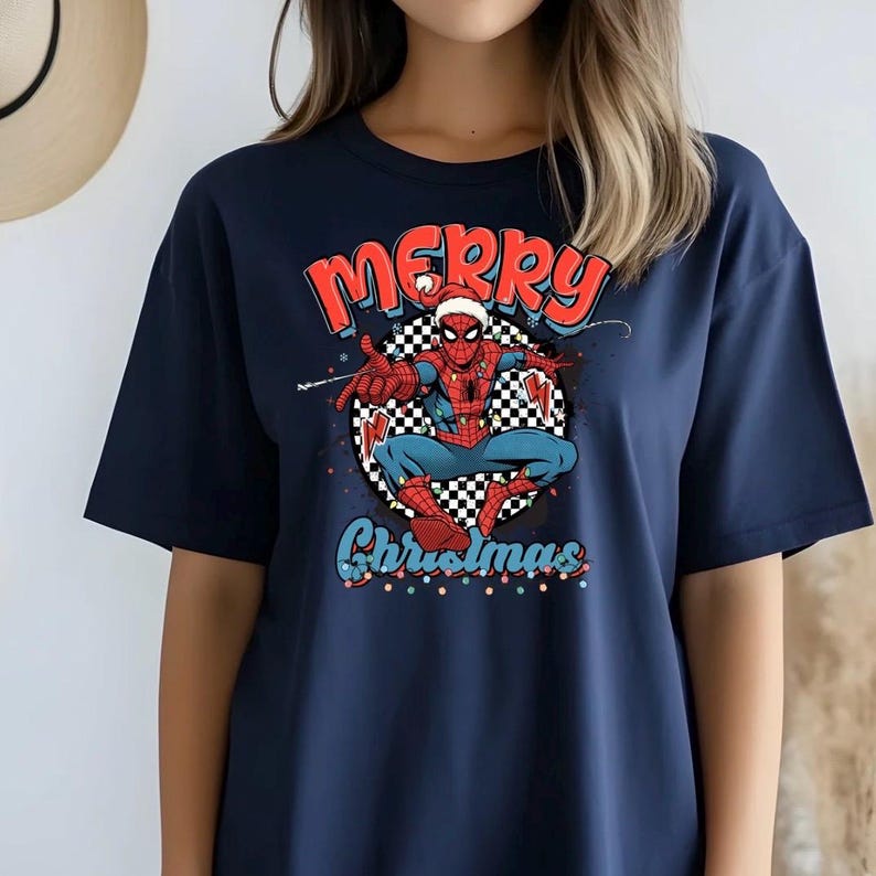 Puede incluir: Camiseta azul marino con un dise&ntilde;o de Spider-Man con gorro de Pap&aacute; Noel y las palabras "Merry Christmas" en rojo y azul. El superh&eacute;roe est&aacute; sobre un fondo a cuadros blanco y negro.